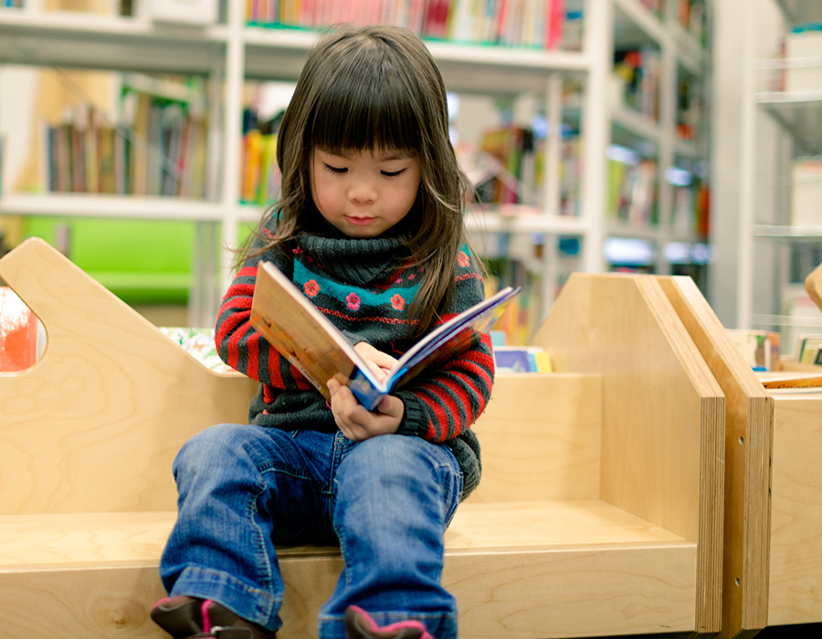 Small asian child reading children's book on a wood library bench